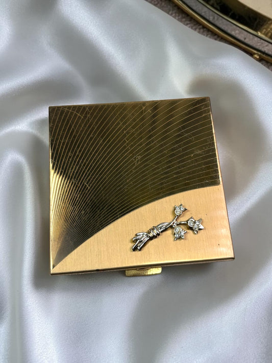 1950s Volupté gold-tone powder compact with engraved starburst design and rhinestone floral accent.