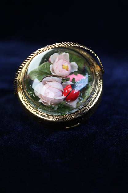 Gold-tone compact with a clear dome showcasing preserved pink flowers and a ladybug.