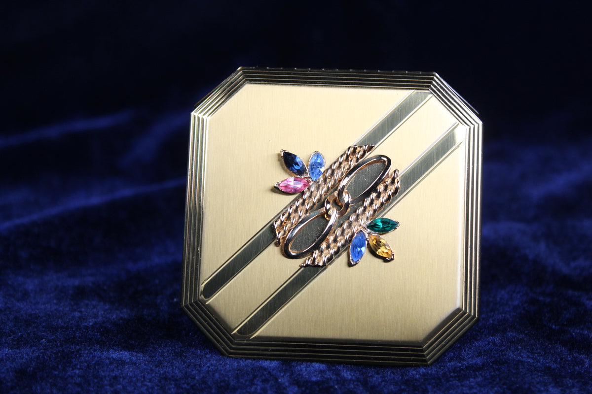 1950s Volupte octagonal gold-tone compact with a rhinestone clasp and decorative diagonal lines.
