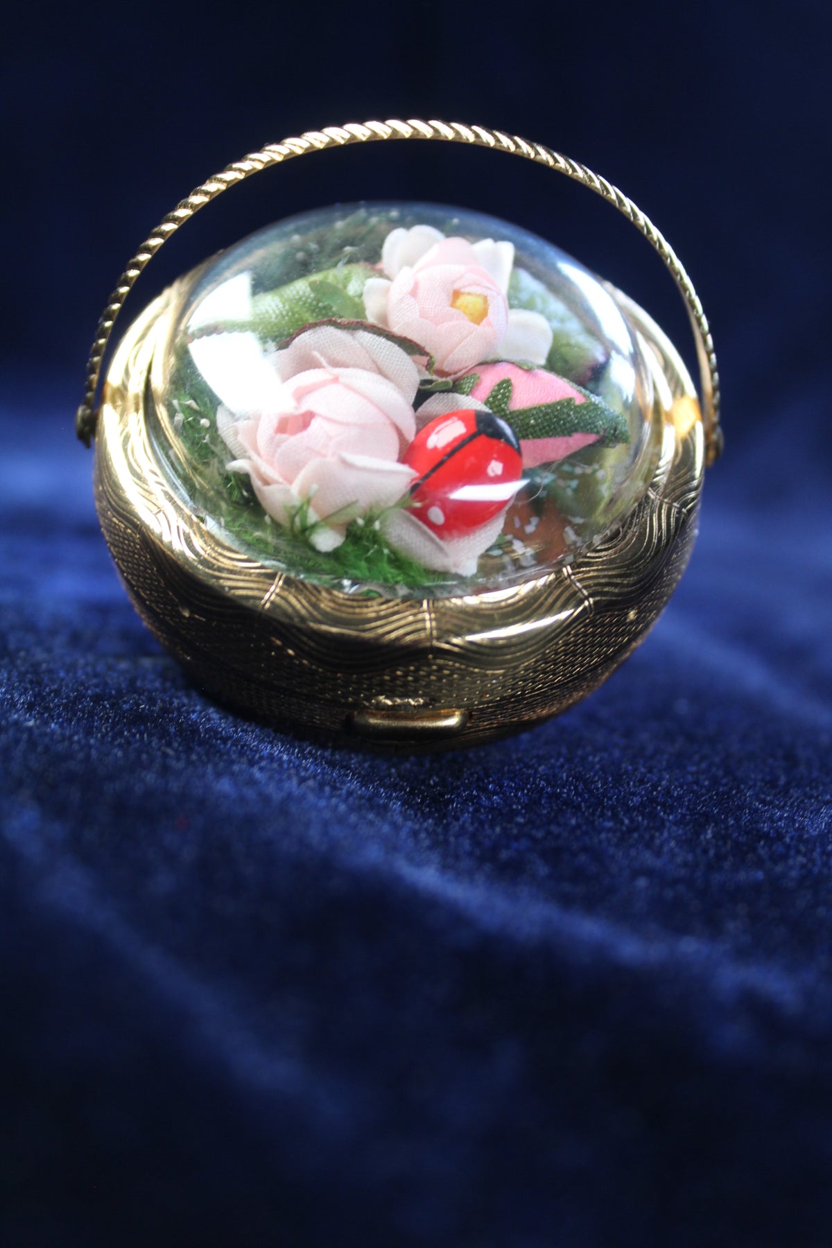 Gold-tone basket compact with preserved pink flowers and a ladybug under a glass dome.