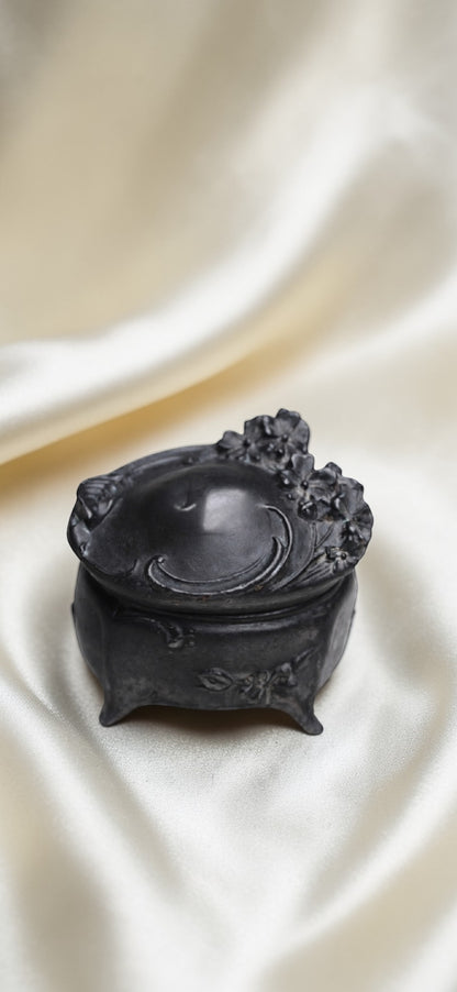 Black metal Art Nouveau trinket box with floral motifs and ornate feet on a cream satin background.