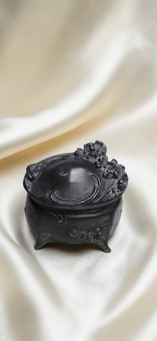 Black metal Art Nouveau trinket box with floral motifs and ornate feet on a cream satin background.