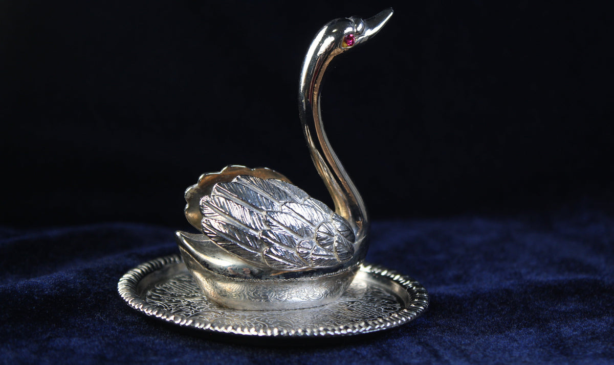 Silver-plated swan ring holder with a purple gemstone eye, resting on an ornate base plate.