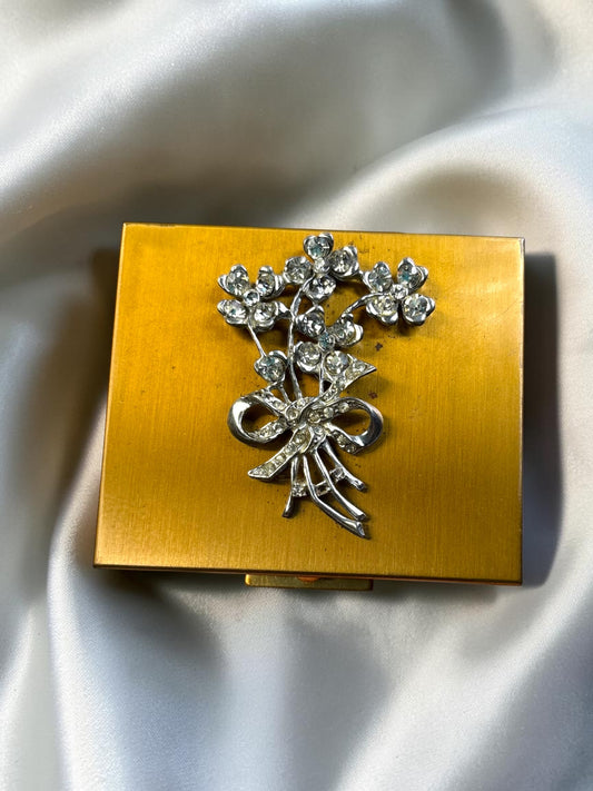 Gold tone powder compact with a rhinestone floral bouquet and bow embellishment.