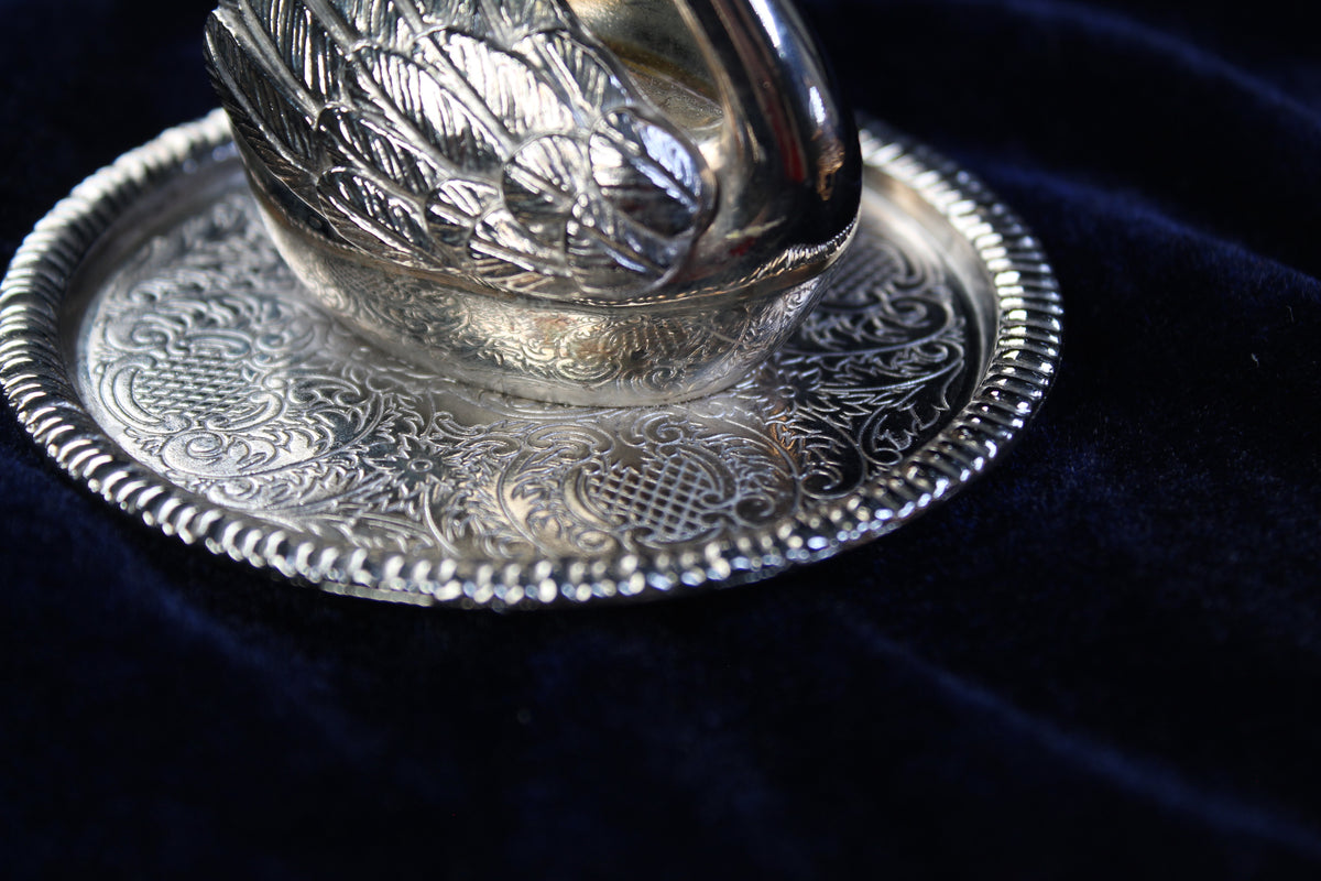 Silver-plated swan ring holder with ornate engraved base on dark blue velvet.