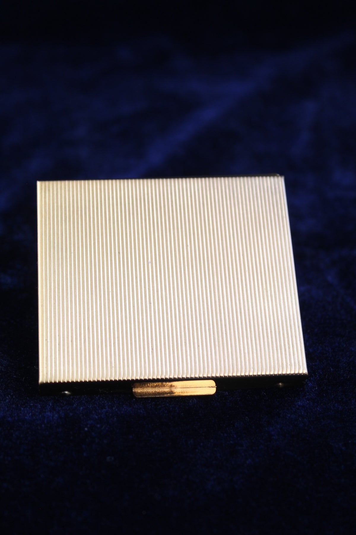 Gold-toned ribbed powder compact with a gold clasp on a dark blue velvet surface.