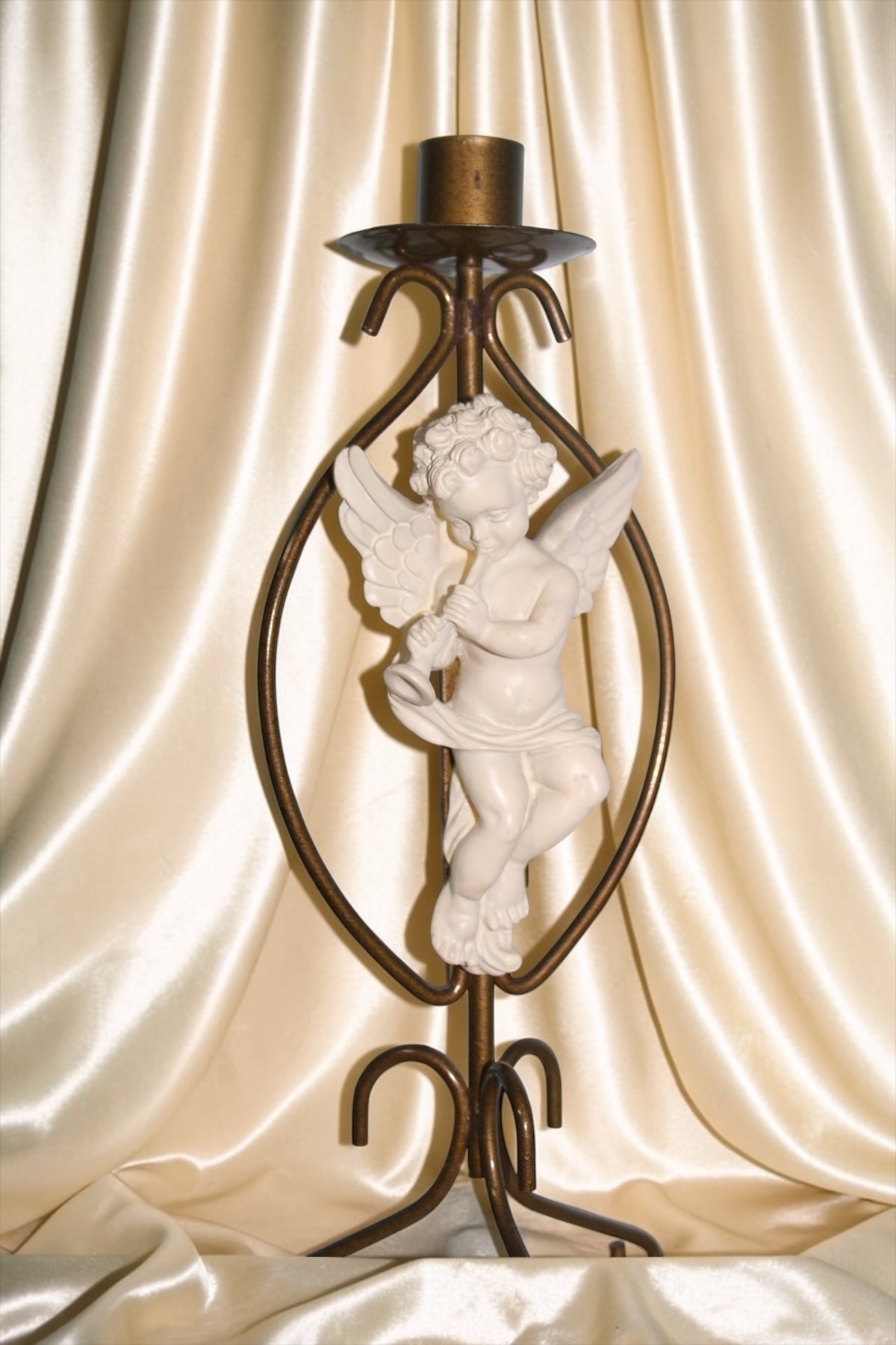Brass candle holder with a white cherub figurine playing a horn, set against a cream satin backdrop.