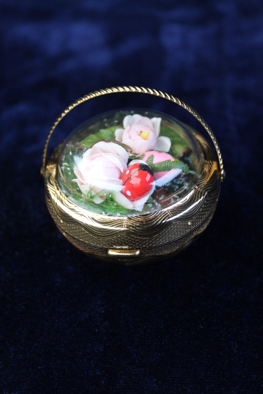 Gold-tone basket compact with preserved pink roses and a ladybug under a glass dome.