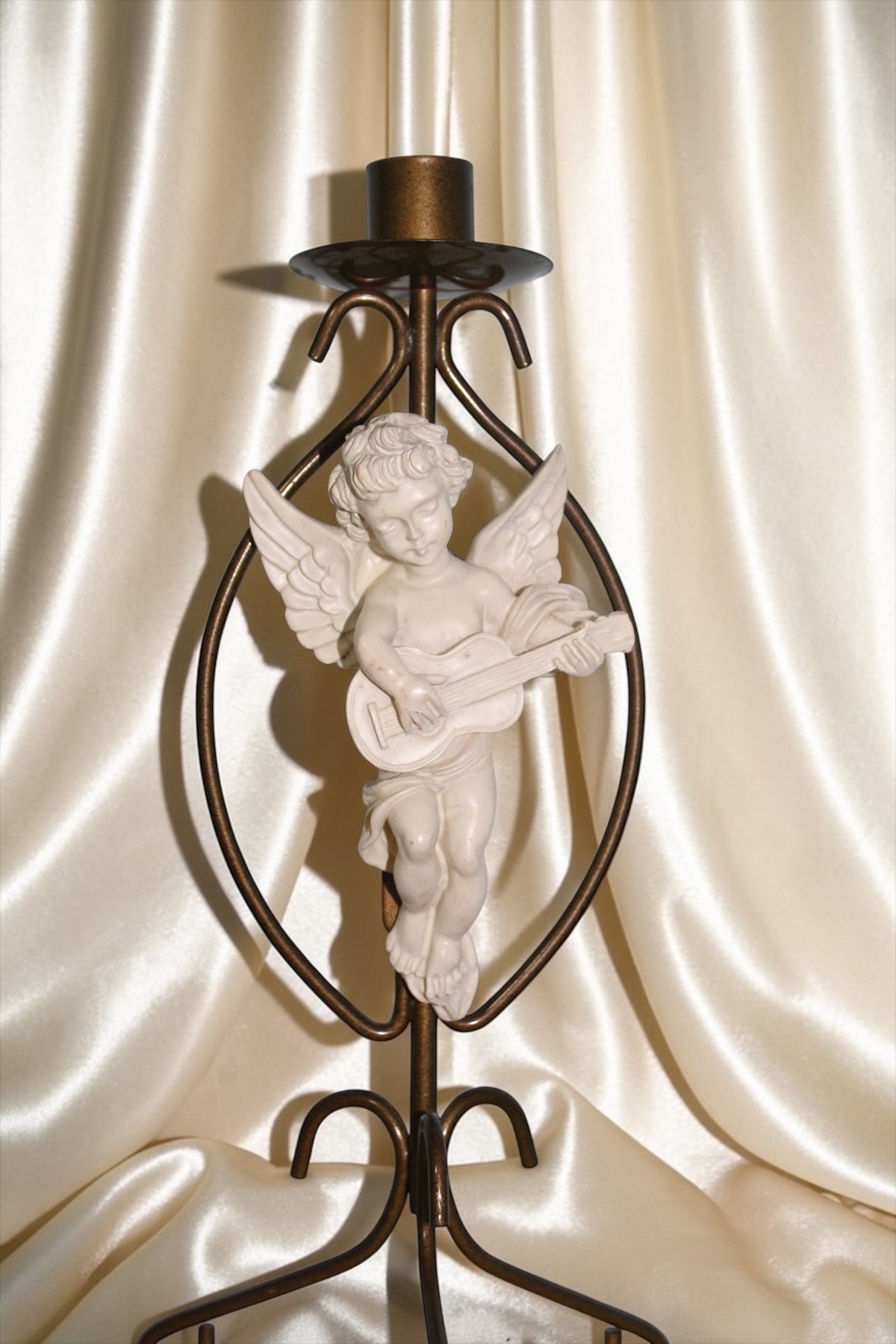 Brass candle holder with a white cherub playing a guitar, set against a cream fabric background.