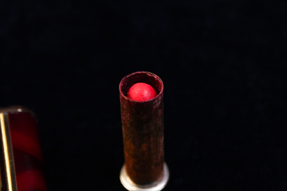 Close-up of a vintage red lipstick bullet inside a tarnished metal tube, against a black background.