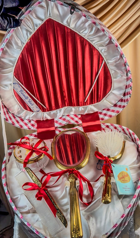 Heart-shaped vanity set with red satin lining, featuring a gilt mirror, hairbrush, comb, and powder jar.