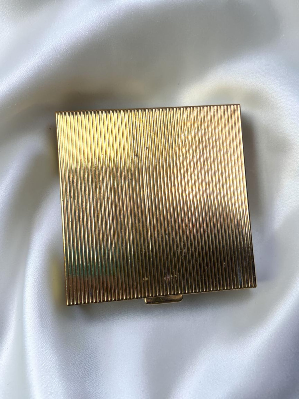 Gold tone ribbed powder compact from the 1950s on a white satin background.