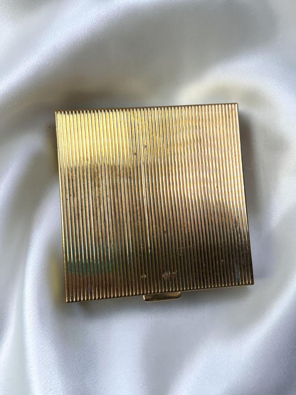 Gold tone ribbed powder compact from the 1950s on a white satin background.