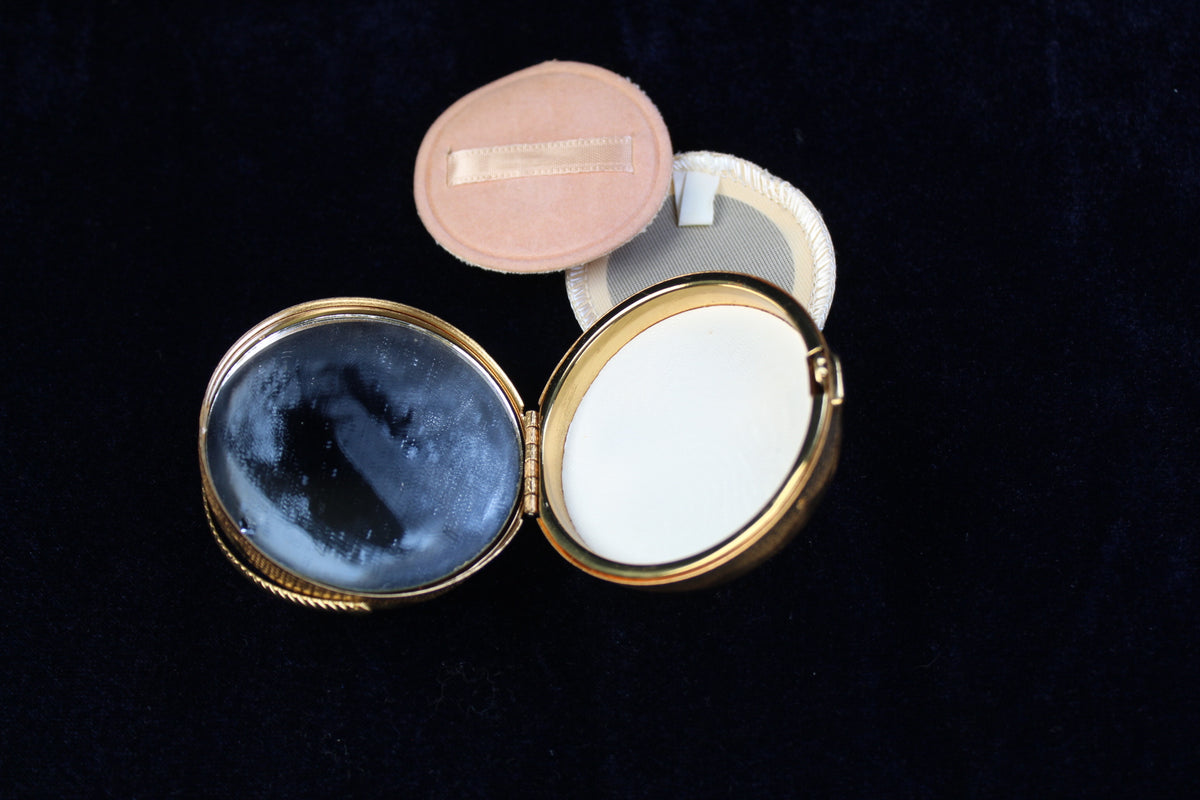 Gold-tone oval compact open to reveal a mirror and white powder, with two powder puffs beside it.
