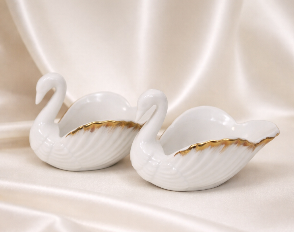 Pair of white porcelain swan figurines with gold trim, resting on a satin cloth.