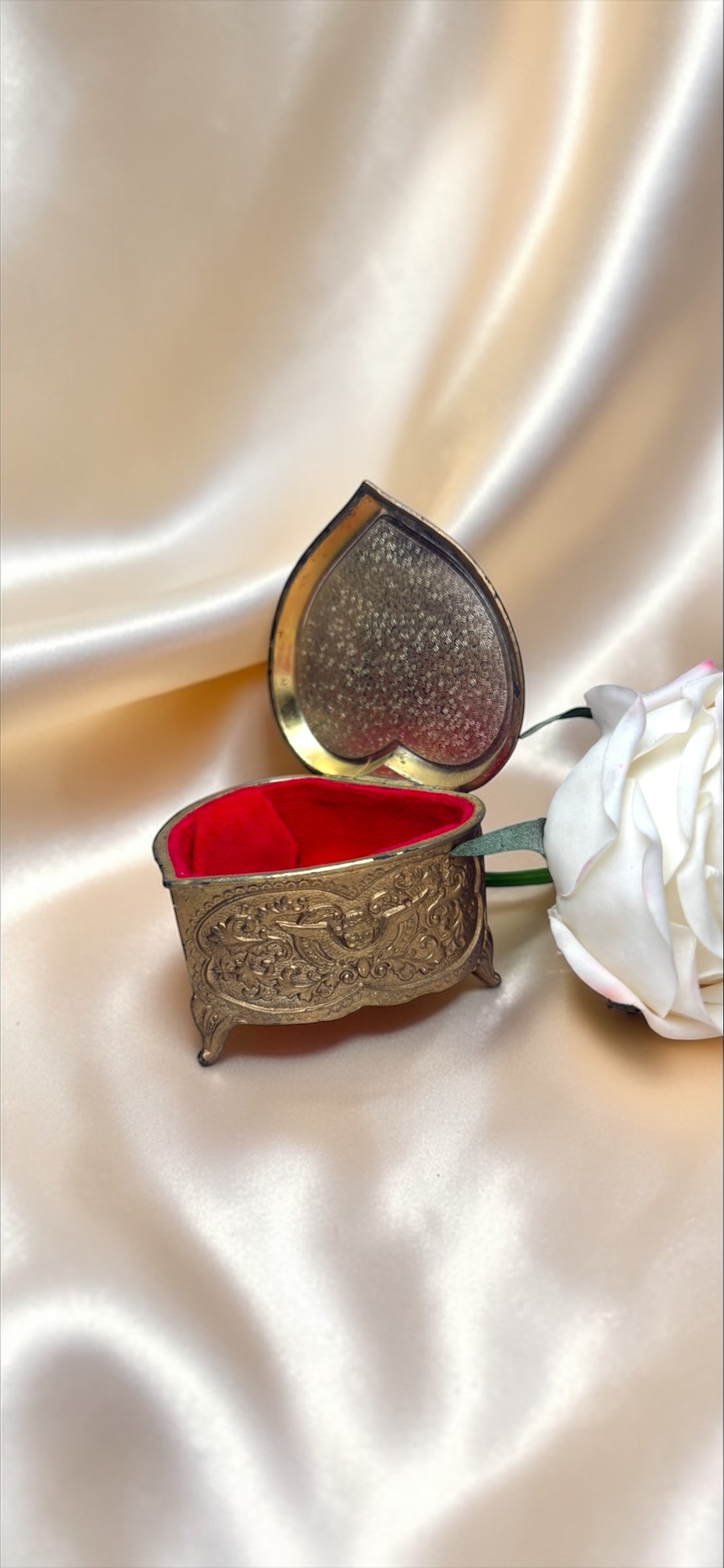 Vintage heart-shaped brass jewelry box with ornate Victorian detailing and a red velvet interior, next to a white rose.