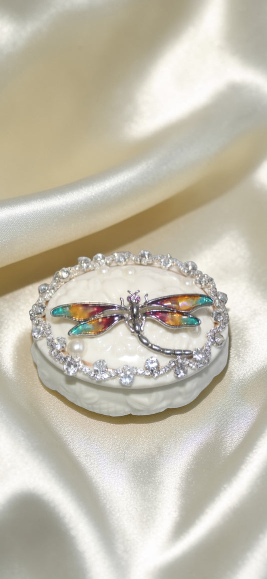 White oval trinket box with a colorful dragonfly brooch and crystal trim on satin.