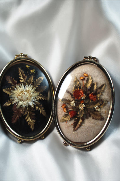 Two oval vintage pressed flower art frames with gold trim. One has white flowers on black, the other has orange flowers on beige.