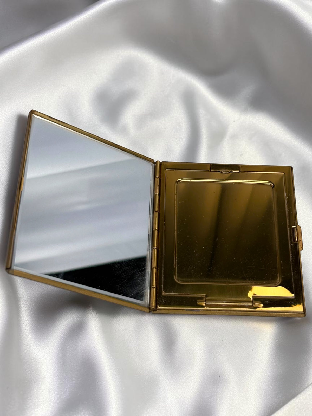 Open gold compact with a mirror on the left and a powder compartment on the right, on a white satin background.
