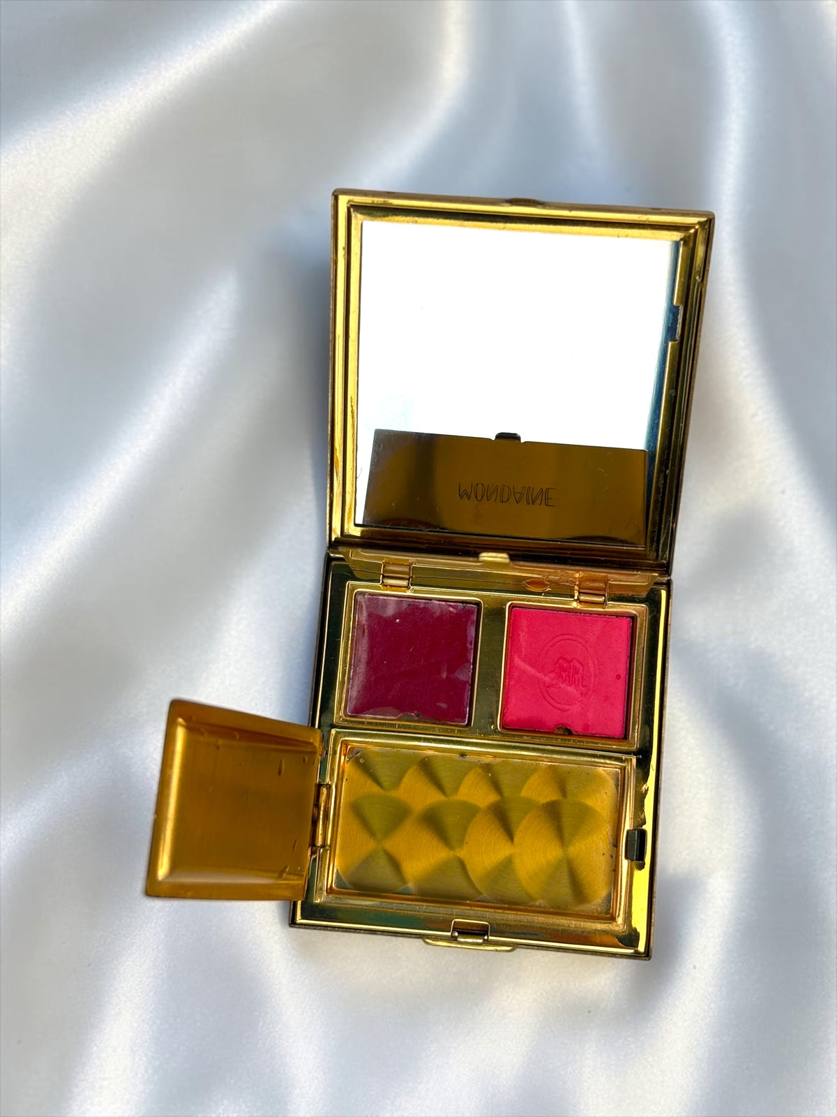 Gold compact with two shades of rouge and a mirror, open on a white satin background.