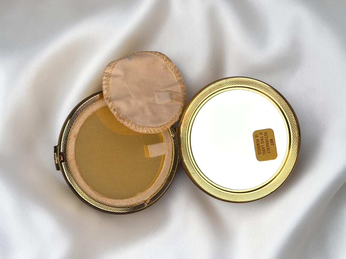 Gold-tone vanity compact open, showing powder puff and mirror with a 'Not Returnable' sticker.